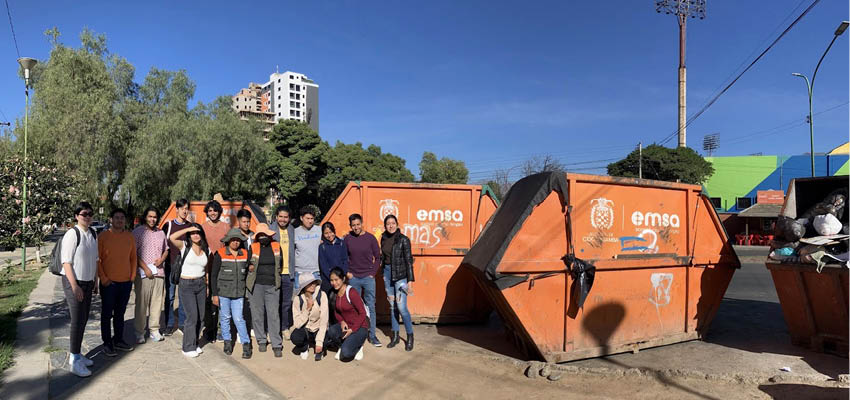 A group of 16 people squinting in the sun in front of two large organge dumpsters.