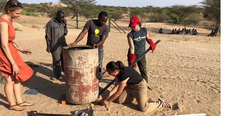 Group of five people around a used metal barrel in a dry sandy area.