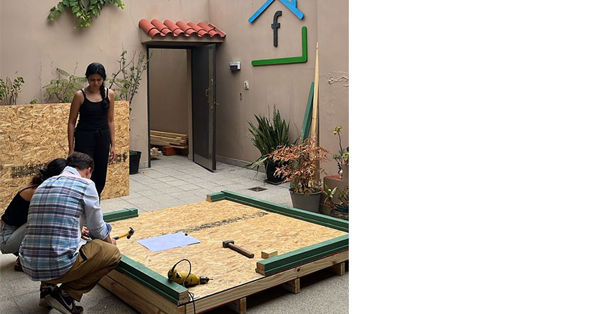 A man crouching in the foreground, buiding a large wooden structure. A woman stands watching.