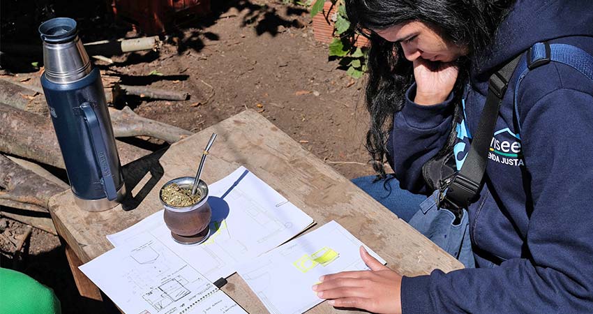 A woman at right, chin in hand, puzzling over documents in front of her on a low, makeshift table outside.