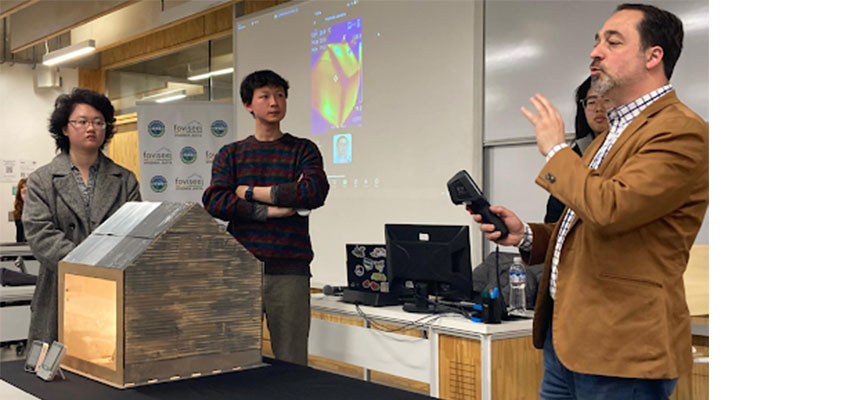 Two students standing left by a model of a house on a table. At right, a man speaks into a microphone.
