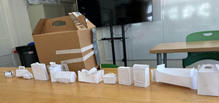 A cardboard box with handles on a table with many small folded white paper boxes in front of it.