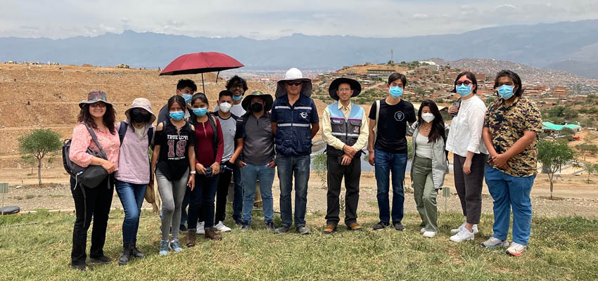 A group of 15 people, most of them with sanitary face masks, on a grassy hill with dry hills, mountains, and clouds behind.