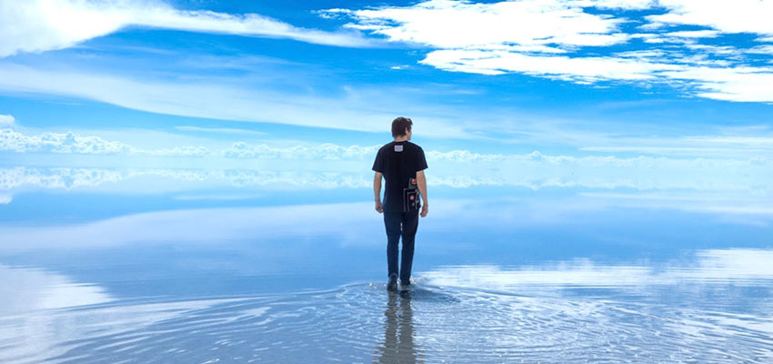 Picture of a man from behind standing outside in shallow water with a large sky and cloudscape in front of him.