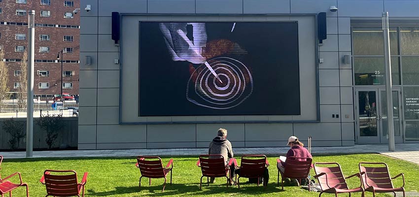 Two people sitting in plastic chairs on a lawn in front of a large screen.