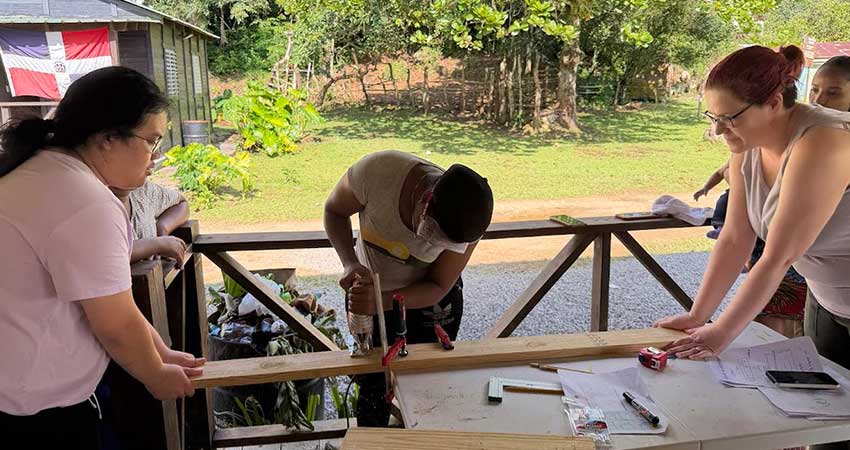 Two women holding either end of a plank of wood while a third, in the middle, saws through the wood.