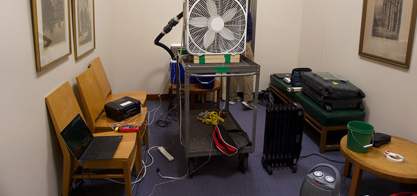 A room with a few arm chairs, a box fan, and a small fan on the floor.