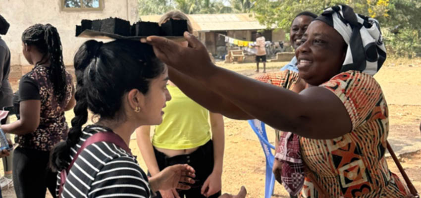 A smiling woman places a small tray of charcoal briquettes on another smiling woman's head.