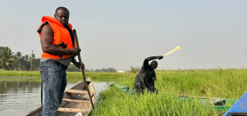 Two mean in canoe like boats, one standing, one chopping water vegetation.