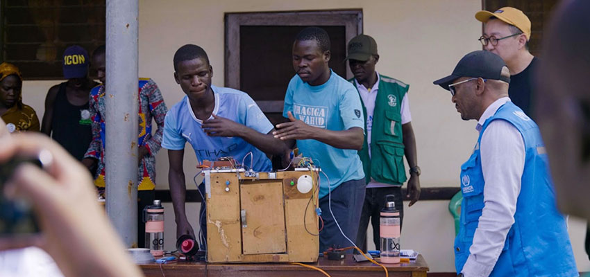 Five men stand behind and around a table wit a large wooden box with wires coming out of it.