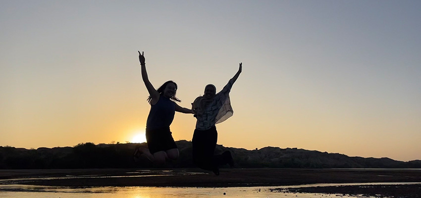 Two women, each with an arm in the air, silhouted against a sunset on a horizon.