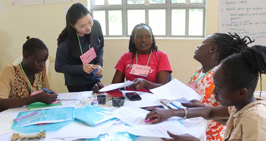 Four women seated at a round table with one additional woman standing. The able is covered in documents.