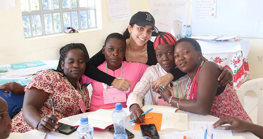 Four women seated at a table, leaning together for the photo. An additional woman crouches behind with arms around one of the seated.