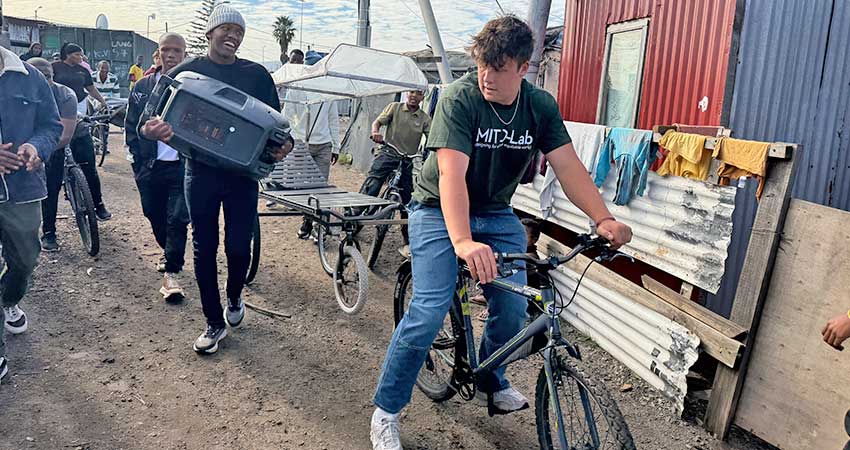 A young man at right, riding a bicycle that is towing a rolling stretcher. A smiling man carrying a box follows at the side.