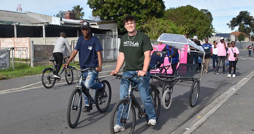 Street scene with man at left astrid a bicycle and man at right astride a bicycle pulling a trailer.