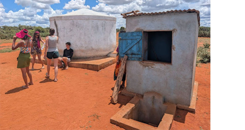 One small round structure at left and one small squarish structure at right on dry red earth. A few people milling around.