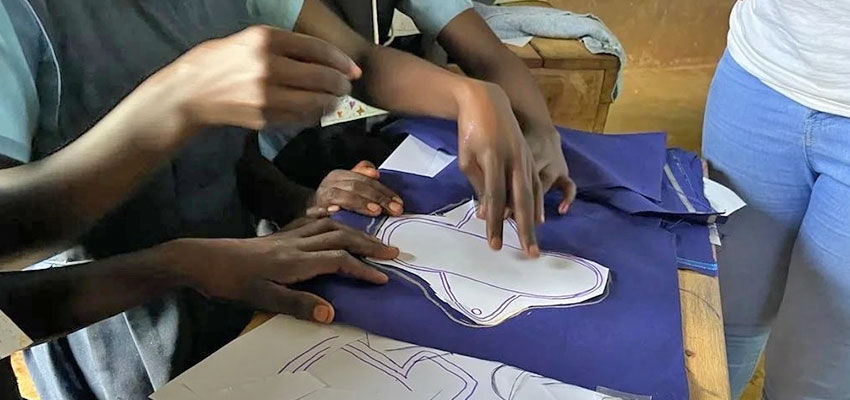 Several hands reaching towards a hand-drawn template of a menstrual pad laid on fabric for cutting and sewing.