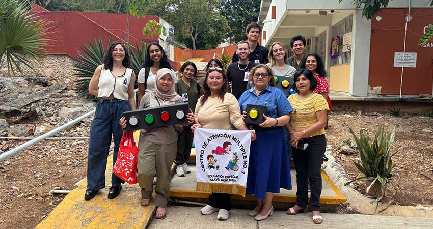 A group of 13 men and women standing on low stairs in front of a one-story buildig, smiling, holding a banner, posting for a group portrait.