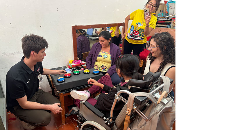 Four people sitting around a small black table, one in wheelchair. A woman is standing at right recording the scene on a cellphone.