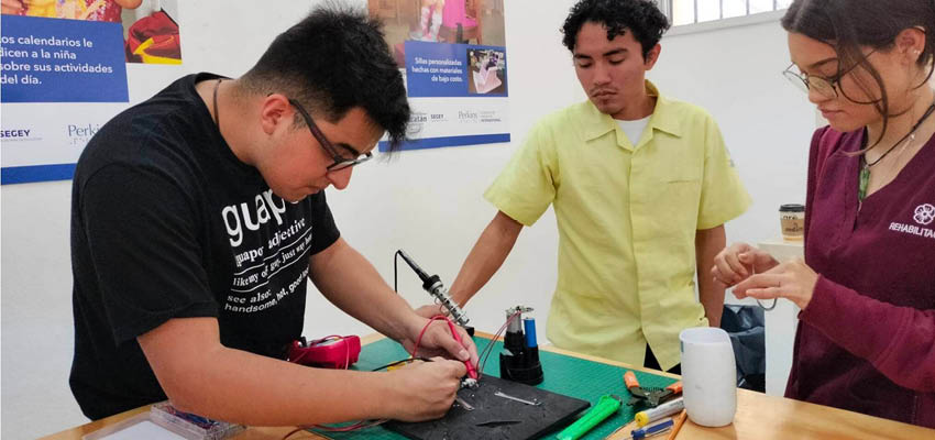 three young adult students working at a workshop table. One student wearing a black shirt and glasses uses a tool to solder wires, while the other two students watch him from the side.