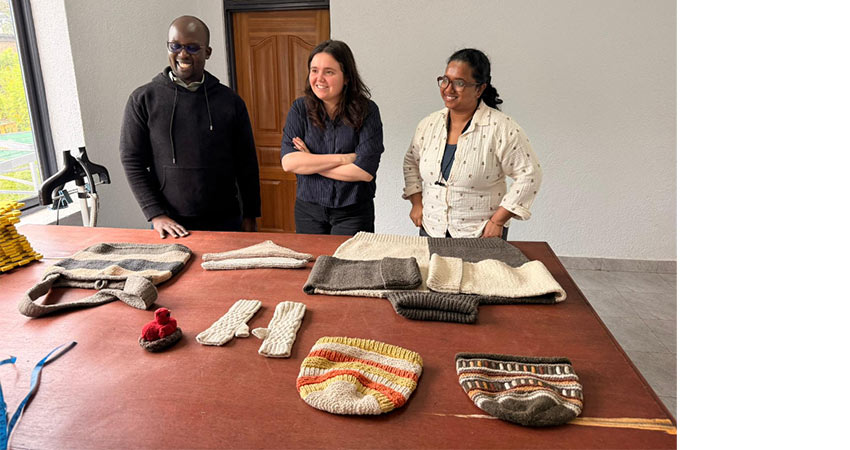 A man and two women stand smiling behind a table on which there are hand-knit sweaters and hats.