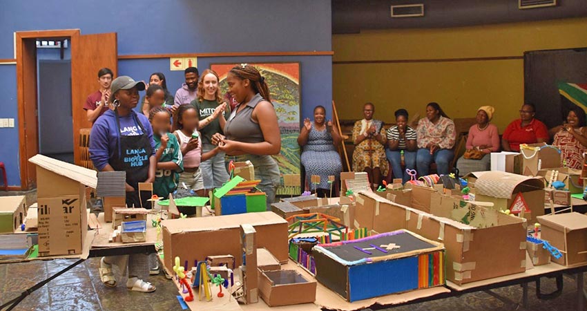 A small group of adults and children standing behind a table covered with cardboard models of buildings. A small group of people is seated in a row in the background at right.
