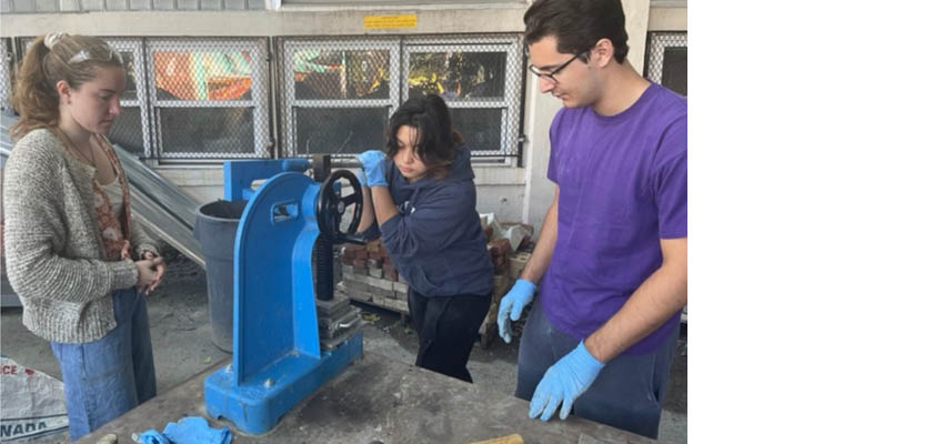 Three young adults at a worktable pulling a lever on a blue mechanical mechanism.