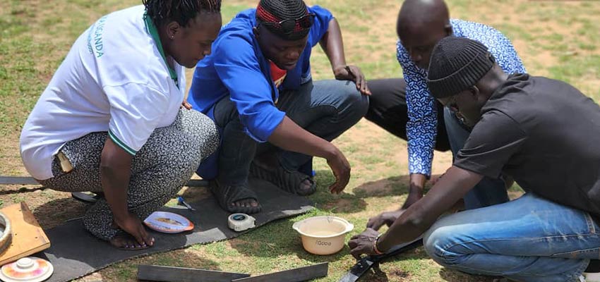 Four men outside crouched over a small plastic bowl and some metal parts.