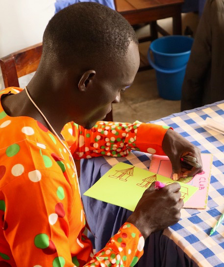 Afrcian man in colorful shirt seated at a table sketching.